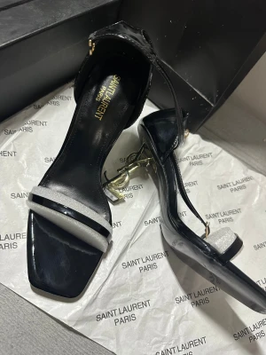 Saint Laurent svarta klackar strl 38 - Säljer ett par lyxiga svarta klackar från Saint Laurent i storlek 38. Skorna har en elegant öppen tå, smala remmar med spänne och ikonisk YSL-logga i guld som klack. Tillverkade i glansigt skinn med silverdetalj framtill. Perfekta för dig som vill sticka ut. Klackarna är aldrig använda! Pris kan diskuteras vid snabb affär☺️