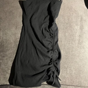 Svart ribbad kort klänning Shein XS - Svart, ribbad kort klänning från Shein i storlek XS. Klänningen är axelbandslös och har snygg snörning på ena sidan som ger en cool rynkad effekt. Perfekt för dig som gillar en tight och trendig look.