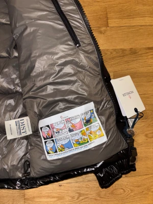 Svart Moncler pufferjacka med huva - Svart glansig pufferjacka från Moncler med huva och dragkedja framtill. Jackan har flera fickor med dragkedja, elastiska muddar och coolt serietryck på insidan. Perfekt för dig som vill ha en snygg och varm jacka med street vibe och exklusiv känsla.