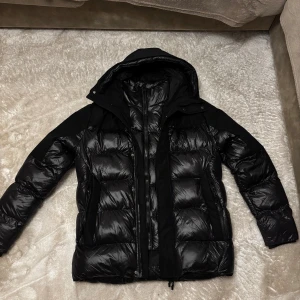 Svart pufferjacka från ZAVETTI CANADA - Säljer en svart pufferjacka från ZAVETTI CANADA med glansig finish och huva. Jackan har flera dragkedjor, praktiska fickor och quiltad design. Perfekt för kalla dagar och har en cool look med tydliga paneler och detaljer.