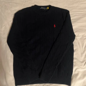 Svart stickad tröja Polo Ralph Lauren - Svart kabelstickad tröja från Polo Ralph Lauren med klassisk rund hals och långärmad passform. Tröjan har röd broderad logga på bröstet och ribbade muddar vid ärmslut och nederkant. Perfekt för dig som gillar stilrena och tidlösa plagg.