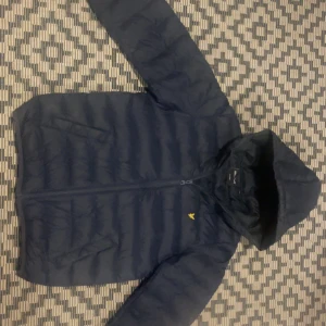 Marinblå pufferjacka Lyle & Scott - Säljer en marinblå pufferjacka från Lyle & Scott med huva och dragkedja. Jackan har ett quiltat mönster och den klassiska gula logon på bröstet. Jackan är andvänd bara en vinter och är i jättefint skick och har inga skador. Köpte den på kidsbrand store och säljer den för att den har blivit liten. Passar nog någon som är 160/172 ungefär.