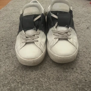 Valentino Garavani vita sneakers - Snygga vita sneakers från Valentino Garavani med blå detalj över snörningen. Skorna har rund tå, platt sula och klassisk snörning. Tillverkade i skinn och levereras med originalkartong. Perfekta för dig som vill ha en stilren och lyxig look.