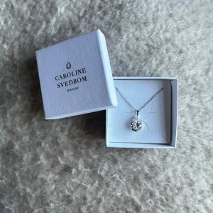 Silver halsband Caroline Svedbom - Stilrent halsband från Caroline Svedbom med en droppformad, glittrig kristall i silverfärg. Kedjan är tunn och elegant, och hänget har en snygg facettslipning som fångar ljuset. Perfekt för dig som gillar klassiska smycken med lite extra bling.