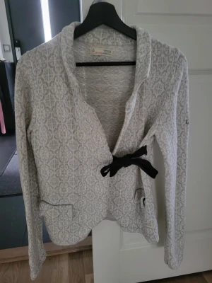 Grå/ vit cardigan jacka Odd Molly med rosett - Snygg grå/vit cardigan jacka från Odd Molly med ljusgrått mönster och svart rosett i midjan. Jackan har lång ärm, krage och broderad kompassdetalj på ena ärmen. Mjukt stickat material och två fickor fram. 