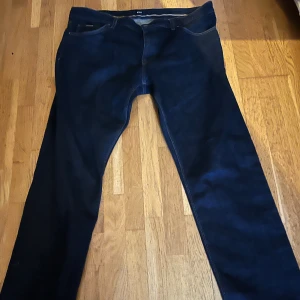 Mörkblå jeans från Hugo Boss - Snygga mörkblå jeans från Hugo Boss med klassisk femficksdesign och brunt läderpatch bak i midjan. Jeansen är tillverkade i stretchigt denim med italiensk kvalitet och har raka ben för en clean look. Perfekta för dig som gillar stilrena och tidlösa jeans.