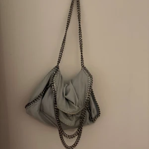 Ljusblå axelväska Stella McCartney - Snygg ljusblå axelväska från Stella McCartney med kedjedetaljer runt kanterna och som axelrem. Väskan har magnetlås, innerfack med dragkedja och ett ljusrosa foder. Perfekt storlek för det viktigaste och har en cool, modern look.