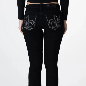 Svarta bootcut jeans Miss Me 28 - Svarta jeans från Miss Me med bootcut passform och låg midja. Bakfickorna har stora silverfärgade paljettbroderier i form av fleur-de-lis. Jeansen har klassisk femficksdesign, snygga detaljer och är tillverkade i stretchig bomullsmix. Storlek W28 L32