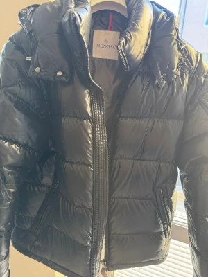 Svart Moncler jacka - Säljer en svart pufferjacka från Moncler med glansig finish, klassisk broderad logga på ärmen och dragkedja framtill. Jackan har hög krage, tryckknappar och två fickor med dragkedja. Insidan har Monclers ikoniska serietidningsetikett. Storlek 2 vilket motsvarar Medium. 7000kr 