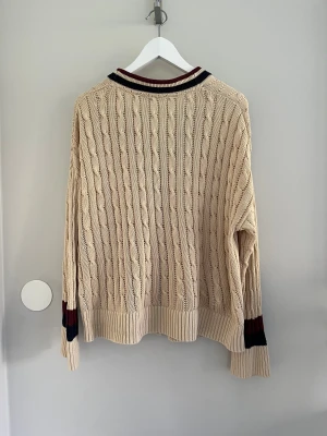Beige kabelstickad kofta Tommy Hilfiger - Snygg beige kabelstickad kofta från Tommy Hilfiger i storlek L/XL. Koftan har ribbade muddar, två fickor fram och kontrastfärgade detaljer i vinrött och marinblått vid krage och ärmslut. Tillverkad i mjuk bomull, perfekt för lager-på-lager under höst och vinter.