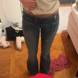Blå bootcut jeans med slitningar - Säljer ett par blå bootcut jeans med låg midja och snygga slitningar på knäna. Jeansen har två knappar i midjan och klassiska fickor fram. Perfekt för dig som gillar en avslappnad och trendig stil. Från Gina young, har tyvärr en liten mens fläck på insidan av jeansen som inte alls syns utanpå