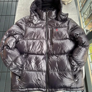 Svart pufferjacka från Polo Ralph Lauren - Svart pufferjacka från Polo Ralph Lauren med glansig finish, huva och flera dragkedjor. Jackan har en röd Polo-logga på bröstet, quiltad design och praktiska fickor med dragkedja. Perfekt för kalla dagar och har en riktigt clean look.