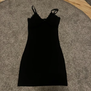 Svart spetsklänning från H&M  - Supersnygg svart kortklänning från H&M i ribbad trikå med smala axelband och spetsdetaljer vid urringningen. Figurnära passform och liten rosett framtill. Storlek xs