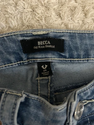 True Religion Becca Bootcut Jeans - Snygga ljusblå jeans från True Religion, modell Becca Mid Rise Bootcut. Klassisk femficksdesign med unika ficklock och silverfärgade knappar. Bootcut-ben och midwaist passform, tillverkade i mjukt jeanstyg med coola detaljer och kontrastsömmar.