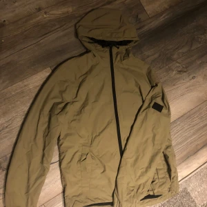 Grön vindjacka med huva - Snygg grön vindjacka med huva och hel dragkedja framtill. Jackan har två fickor med lock och en diskret svart patch på ena ärmen. Perfekt för blåsiga dagar och enkel att packa ner. Lätt och smidig design med lång ärm