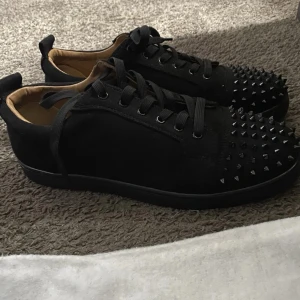 Svarta Christian Loubotins Med Spikar - Säljer ett par riktigt feta svarta sneakers från Christian Louboutin med coola svarta nitar på tån och klassisk röd sula. Skorna har snörning och är gjorda i mocka med skinnfoder. Perfekt för dig som vill sticka ut med en edgy och lyxig stil.