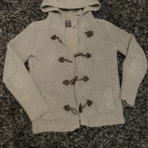 Grå stickad kofta med huva och träknappar - Säljer en grå stickad kofta från Denim med huva och snygga träknappar framtill. Koftan har grov ribbstickning, två fickor och långa ärmar. Perfekt för dig som gillar chill och stilren look.