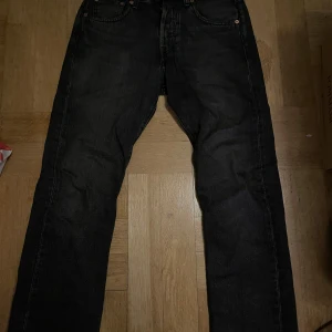 Levi's 501 svarta jeans W27 L28 - Svarta Levi's 501 jeans med klassisk rak passform och fem fickor. Jeansen har knappgylf och är tillverkade i slitstarkt bomullsmaterial. Perfekt för dig som gillar tidlös stil och vill ha ett par jeans som funkar till allt. Storlek W27 L28.