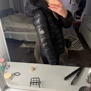 Moncler bady  - Säljer en svart dunjacka från Moncler med glansig finish och huva. Jackan har Moncler-logga på ärmen, dragkedja framtill och är quiltad för extra värme. Perfekt för kalla dagar och har en snygg, modern look.