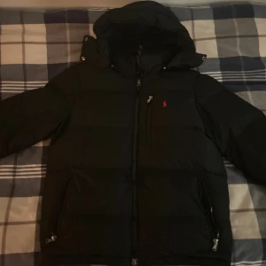 Svart pufferjacka från Polo Ralph Lauren - Svart pufferjacka från Polo Ralph Lauren med huva och dragkedja framtill. Jackan har en röd Polo-logga på bröstet, två sidofickor med dragkedja och en bröstficka med dragkedja. Perfekt för kalla dagar och har en clean, stilren look.