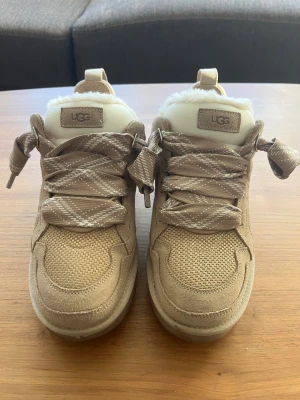 Beige sneakers från UGG med snörning - Säljer ett par beige sneakers från UGG med breda, mönstrade snören och fluffigt foder. Skorna har en rund tå, platt sula och är tillverkade i mocka och syntet med stickade detaljer. Perfekta för dig som gillar en cozy och snygg stil. Använt några gånger, men ej några störrre synliga defekter 