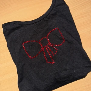 Svart topp med röd rosett i paljetter - Svart topp med en stor rosett framtill, dekorerad med röda paljetter. Rund halsringning och mjukt. Tröjan är i grund från lager 157, går att tvätta OBS! Bara i 30 grader, ingen torktumling 