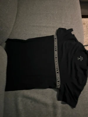 Svart topp från Armani Exchange XS - Svart kortärmad topp från Armani Exchange i slim fit. Snygg detalj med ett band över bröstet där det står 'Armani Exchange'. Mjuk och skön bomullskvalitet, perfekt för dig som gillar stilrena och moderna plagg med logga.