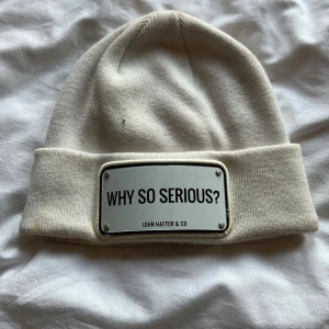 Vit mössa John Hatter & Co - Snygg vit mössa från John Hatter & Co med uppvikt kant och en metallbricka framtill med texten 'WHY SO SERIOUS?'. Tillverkad i mjukt material som sitter skönt på huvudet. Perfekt accessoar för att sticka ut i mängden.