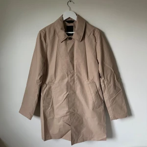 Beige trenchcoat från J.Lindeberg, strl 48 - Snygg beige trenchcoat från J.Lindeberg i stilren design med klassisk krage och knappstängning. Jackan har två fickor fram och är tillverkad i mjuk twill av 100% polyester. Perfekt för dig som gillar minimalistisk och clean stil. 5I13