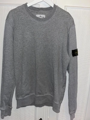 Grå sweatshirt från Stone Island - Säljer en stilren grå sweatshirt från Stone Island med klassisk rund hals och den ikoniska patchen på vänster ärm. Tröjan har ribbade muddar och är tillverkad i mjuk bomull för skön komfort. Säljs pga för liten storlek, använd fåtal gånger. Inköpt för över 2000kr. 