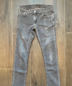 Grå jeans från Nudie Jeans - Snygga grå jeans från Nudie Jeans med klassisk femficksdesign och kontraststickningar. Modellen Long John med låg midja och raka ben. Jeansen har en cool tvättad look och Nudie-loggan på bakfickan. Perfekta för dig som gillar stilrena och moderna jeans.