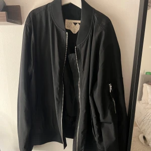 Svart bomberjacka från Junkyard - Svart bomberjacka från Junkyard med klassisk ribbad krage och muddar. Jackan har dragkedja framtill och en ficka med dragkedja på ärmen. Snygg, stilren och passar perfekt till streetwear. Materialet är lätt och känns skönt mot kroppen.