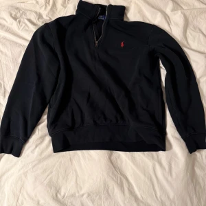 Svart half zip tröja Polo Ralph Lauren - Svart långärmad tröja från Polo Ralph Lauren med half zip och röd broderad logga på bröstet. Tröjan har en hög krage och är gjord i mjuk bomull, perfekt för chill dagar. Klassisk och stilren design som funkar till jeans eller joggers.