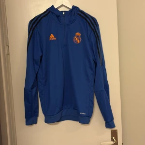 Blå Real Madrid hoodie från Adidas - Blå hoodie från Adidas med Real Madrid-logga i orange på bröstet och svarta ränder längs axlarna. Tröjan har huva, lång ärm och half zip framtill. Materialet är polyester och modellen är normal passform. Perfekt för fotbollsfans som vill ha en sportig look.