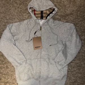 Grå hoodie från Burberry med dragkedja - Snygg ljusgrå hoodie från Burberry med klassiskt rutigt foder i huvan, broderad logga på bröstet och hel dragkedja. Tröjan har snörning i huvan och praktiska fickor framtill. Perfekt för dig som vill ha en stilren och exklusiv look.