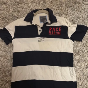 Randig Race Marine skjorta XS - Snygg skjorta från Race Marine i storlek XS med breda marinblå och vita ränder. Skjortan har klassisk krage, korta ärmar och tryckt Race Marine-logga i rött på bröstet. Tillverkad i mjuk bomull, perfekt för en sportig och avslappnad stil.