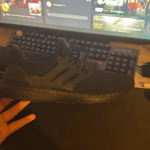 Svarta Adidas Ultraboost sneakers - Säljer ett par svarta Adidas Ultraboost sneakers med stickad ovandel och klassiska tre ränder på sidan. Skorna har platt sula med Boost-dämpning och snörning framtill. Perfekt för dig som gillar stilrena och bekväma sneakers med sportig vibe.