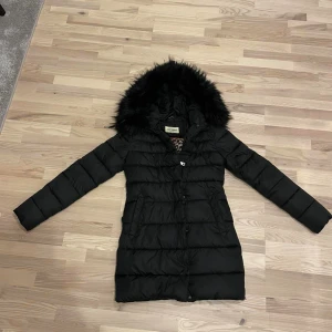 Svart dunjacka med päls från Roco Baroco - Snygg svart dunjacka från Roco Baroco med fluffig svart fuskpäls på huvan och leopardmönstrat innerfoder. Jackan är quiltad, har långa ärmar och praktiska fickor framtill. Perfekt för kalla dagar och har en stilren look.