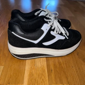Svarta och vita sneakers med snörning - Snygga svarta sneakers med vita detaljer och vit sula. Skorna har klassisk snörning och en rund tå. Ovandelen är i mesh och syntet med kontrasterande vita partier på sidorna. Perfekta för dig som gillar stilrena och sportiga skor.