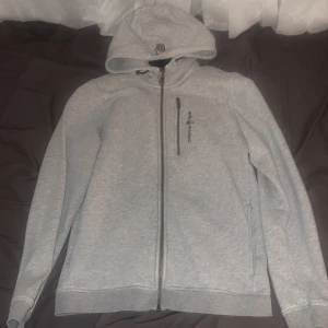 Ljusgrå hoodie från Sail Racing - Säljer en ljusgrå hoodie från Sail Racing med dragkedja hela vägen med huva