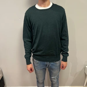 Mörkgrön stickad tröja från Riley - En stilren mörkgrön stickad tröja från Riley med rund halsringning och långa ärmar. Tröjan har ribbade muddar vid ärmslut och nederkant, vilket ger en snygg passform. Perfekt att bära över en t-shirt för en clean look. Hör av er om ni har funderingar eller frågor!