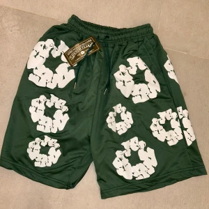 Gröna shorts från Denim Tears med tryck - Säljer ett par gröna shorts från Denim Tears med unikt vitt tryck över hela plagget. Modellen har elastisk midja med snörning och en bakficka. Materialet är mjukt och känns som polyester, perfekt för chill dagar. Designen sticker ut med de grafiska cirklarna.