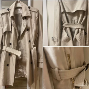 Beige trenchcoat med bälte - Snygg beige trenchcoat med klassisk dubbelknäppning, axelklaffar och bälte i midjan. Jackan har långa ärmar, svarta knappar och detaljer som dragkedja och fickor. Perfekt för dig som gillar stilrena och tidlösa plagg.
