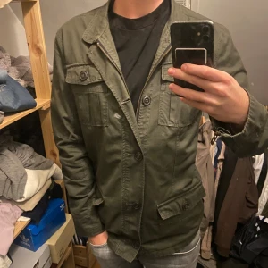 Olivgrön parkajacka med fickor - Säljer en olivgrön parkajacka med flera praktiska fickor och både dragkedja och knappar framtill. Jackan har klassisk krage och är långärmad, perfekt för dig som gillar en clean och funktionell stil. Materialet är bomull och jackan har en loose passform. 