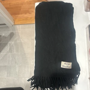 Svart halsduk från Acne Studios - Stilren svart halsduk från Acne Studios med klassiska fransar i kanterna och en diskret logotyp-patch. Halsduken är mjuk och stor. Materialet känns lyxigt (Ull) och passar perfekt till kalla dagar.