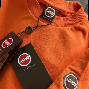 Orange sweatshirt från italienska Colmar NY - Snygg orange sweatshirt från Colmar med rund hals och ribbade muddar. Tröjan har ett stilrent Colmar-märke på ärmen och är tillverkad i mjuk bomull. Perfekt för dig som gillar färg och vill sticka ut lite extra.