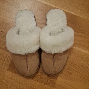 Beige fluffiga tofflor i storlek 37/38 - Mysiga beige tofflor med fluffig vit päls runt kanten och insidan. Utsidan är i mocka och sulan är platt och mönstrad i gummi.