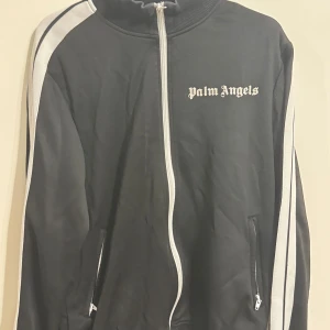 Svart track jacket från Palm Angels - Svart track jacket från Palm Angels med vita ränder längs ärmarna och vit dragkedja framtill. Jackan har ribbad krage och muddar, samt två fickor med dragkedja. Palm Angels-logga broderad på bröstet. Perfekt för en sportig och streetwear-inspirerad look.