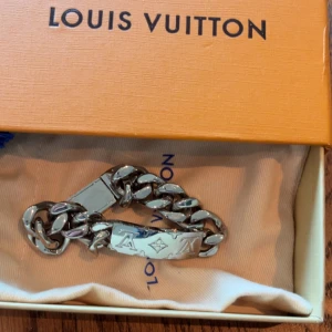 Louis Vuitton silverfärgat armband - Stilrent armband från Louis Vuitton i silverfärgad metall med grov kedja och klassisk LV-logga ingraverad på plattan. Kommer med original ask och dustbag. Perfekt accessoar för dig som gillar exklusiva detaljer och vill sticka ut.