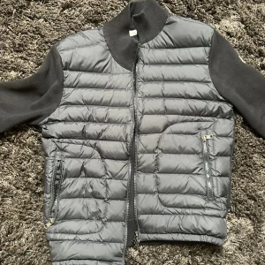 Moncler Cardigan - Snygg svart dunjacka från Moncler med quiltad front och stickade svarta ärmar. Jackan har två dragkedjeförsedda fickor, ribbad krage och Moncler-logga på ärmen. Perfekt för dig som gillar stilren design och exklusiva märken.